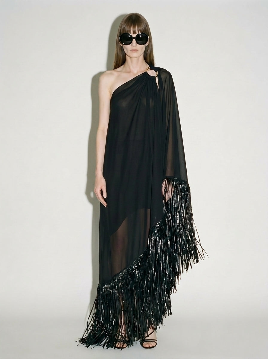 Woman wearing black sheer silk one-shoulder maxi dress with asymmetric fringe trim and gold-tone ring detail.